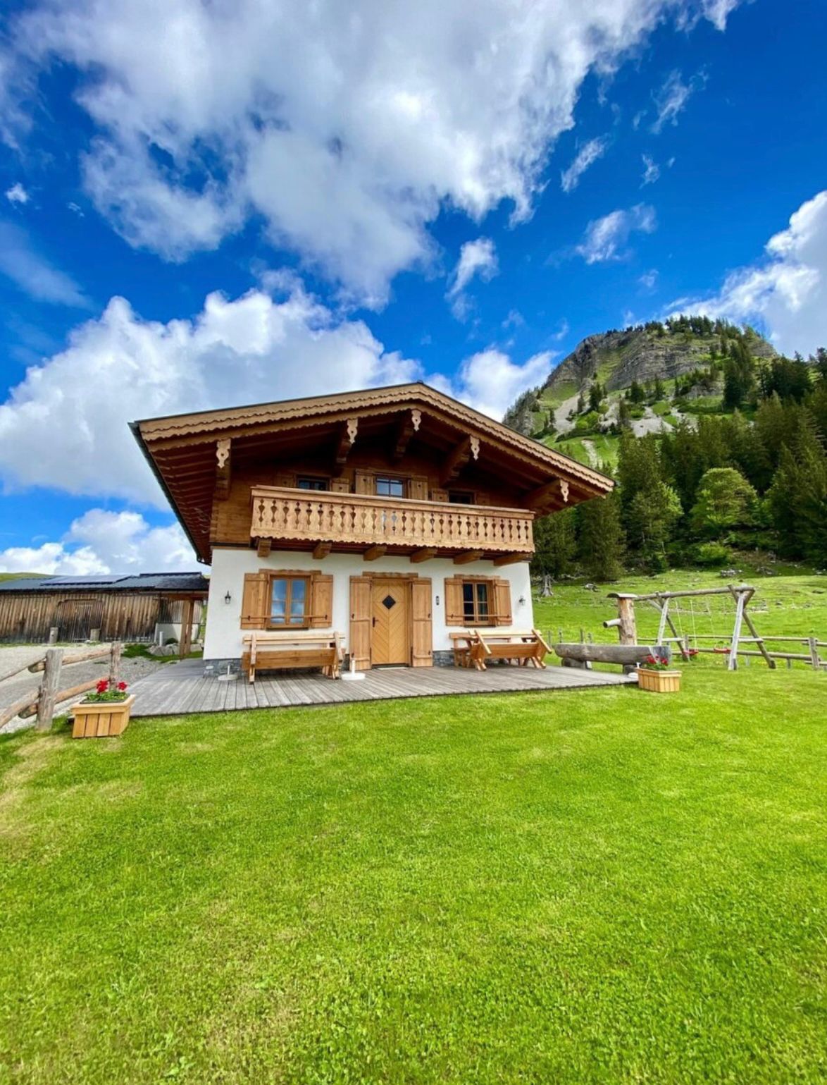 Holzchalet mit Balkon auf gr&uuml;nem Gras, Berge und B&auml;ume im Hintergrund unter einem blauen Himmel mit Wolken.