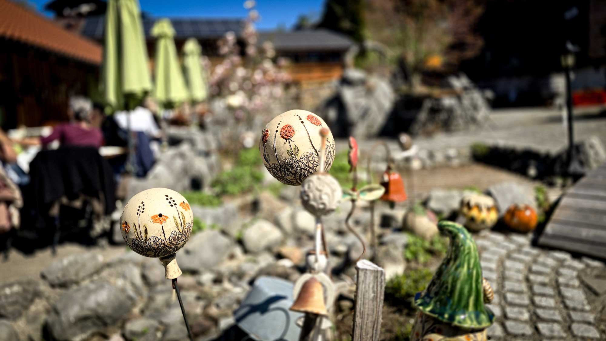 Dekorativer Gartenschmuck aus Keramik in Kathis Blumencaf&eacute; in einer sonnigen Sitzecke mit Steinen und Sonnenschirmen.