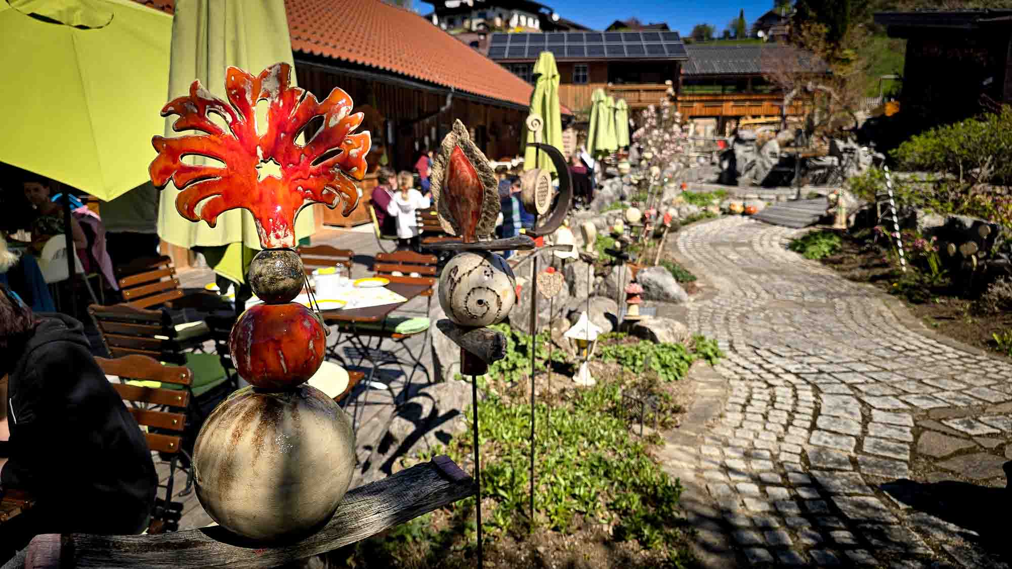 Bunte Keramik-Gartenskulpturen in Kathis Blumencaf&eacute; neben einem Kopfsteinpflasterweg und gr&uuml;nen Sonnenschirmen.