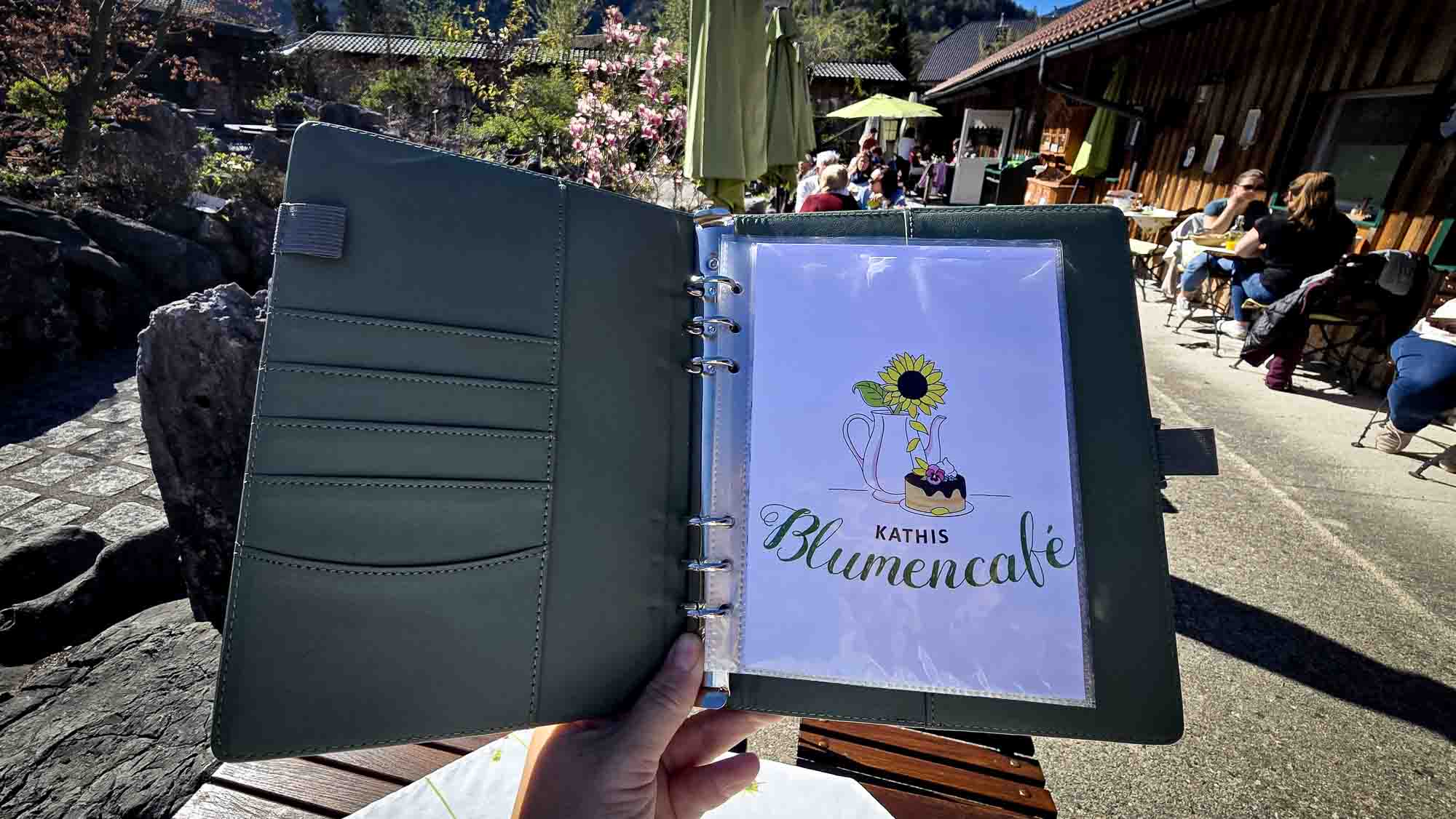 Eine Hand h&auml;lt eine Speisekarte von Kathis Blumencaf&eacute; in einem Stra&szlig;encaf&eacute;, in dem die Menschen die Sonne genie&szlig;en.