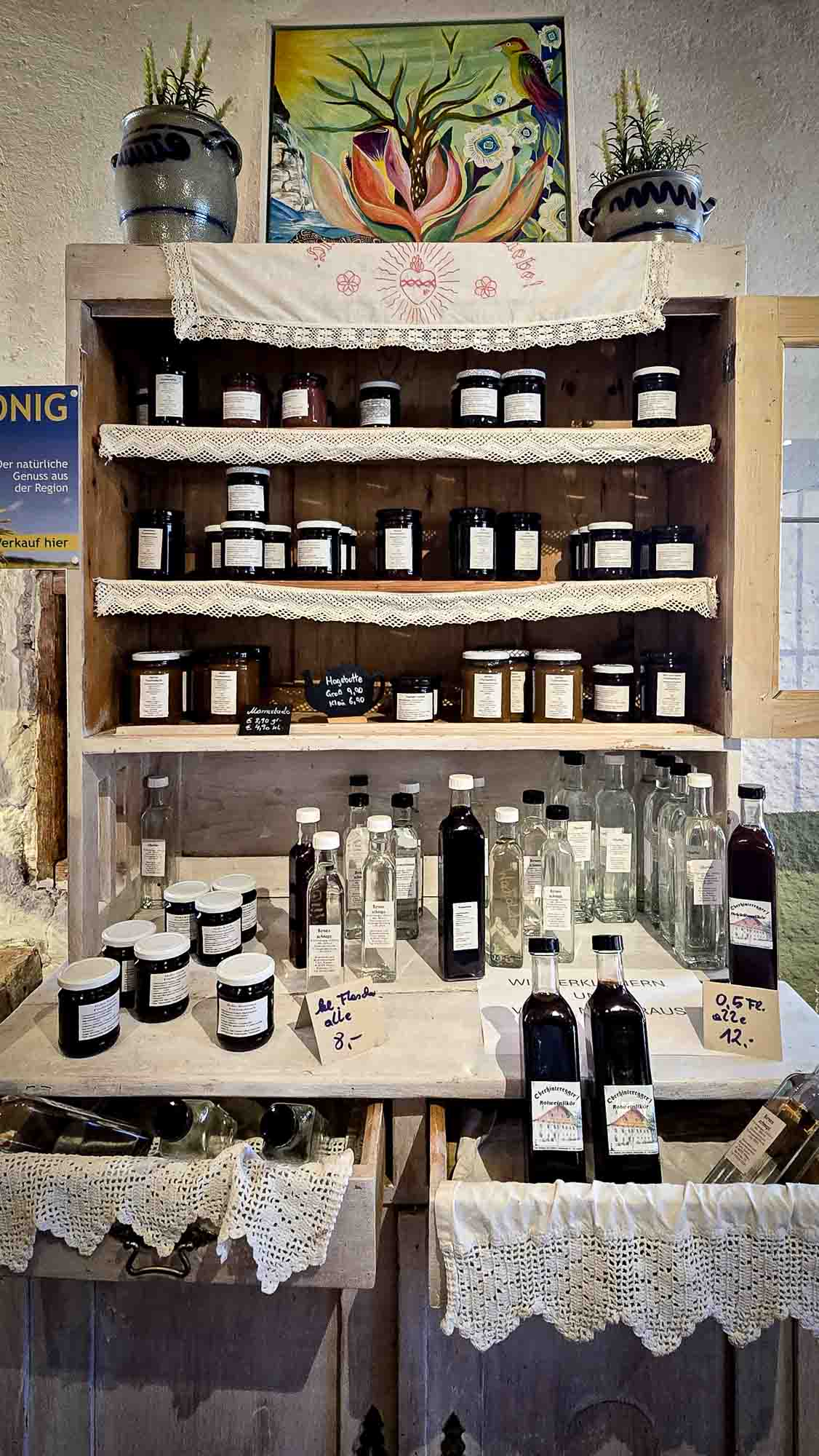 Holzregal in Kathis Blumencaf&eacute; mit Gl&auml;sern, Flaschen und Spitzendeckchen f&uuml;r selbstgemachte Konfit&uuml;ren und Lik&ouml;re.