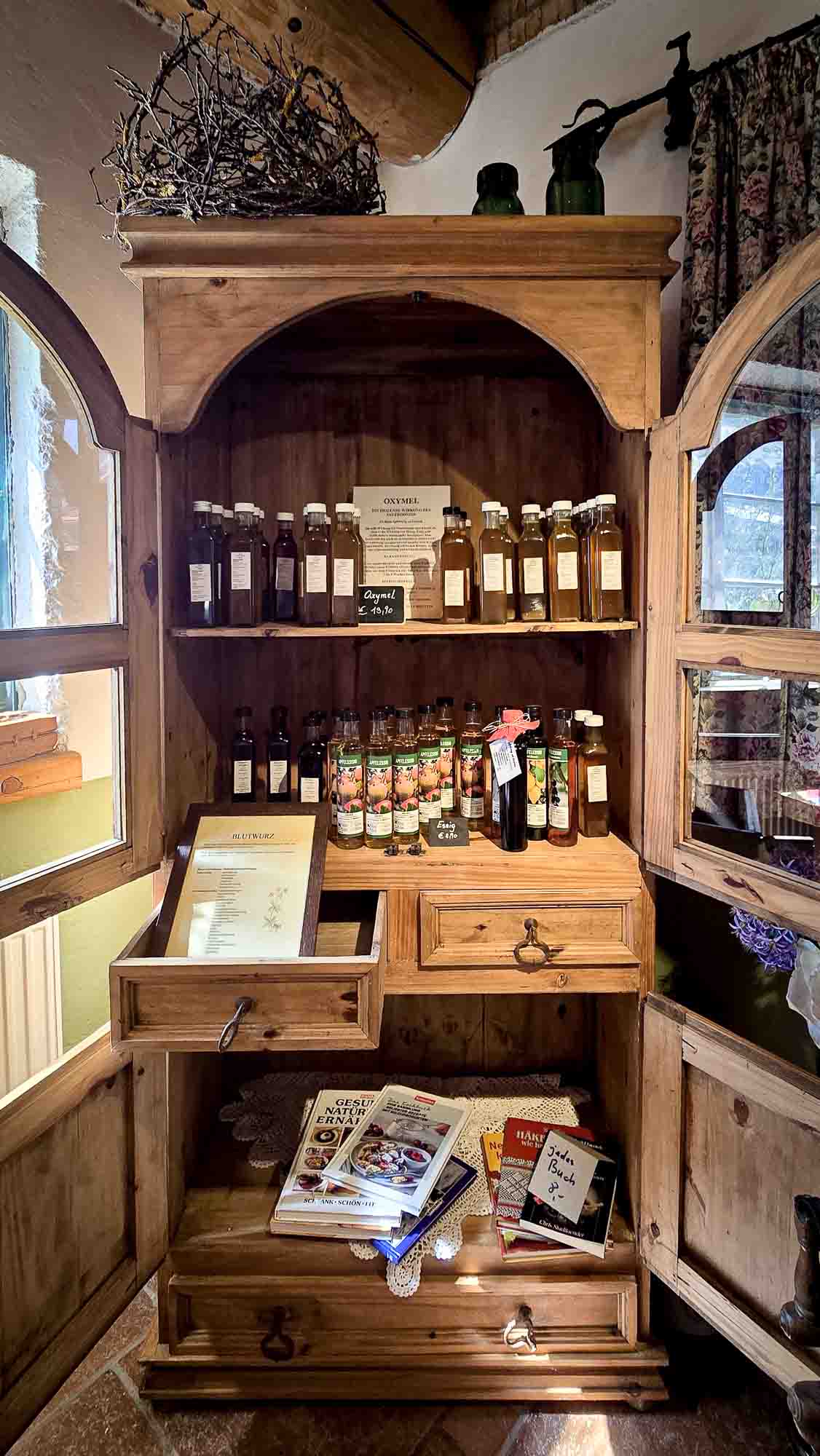 Holzschrank in Kathis Blumencaf&eacute; mit Honiggl&auml;sern, B&uuml;chern, einer Speisekarte auf Regalen und einer offenen Schublade.