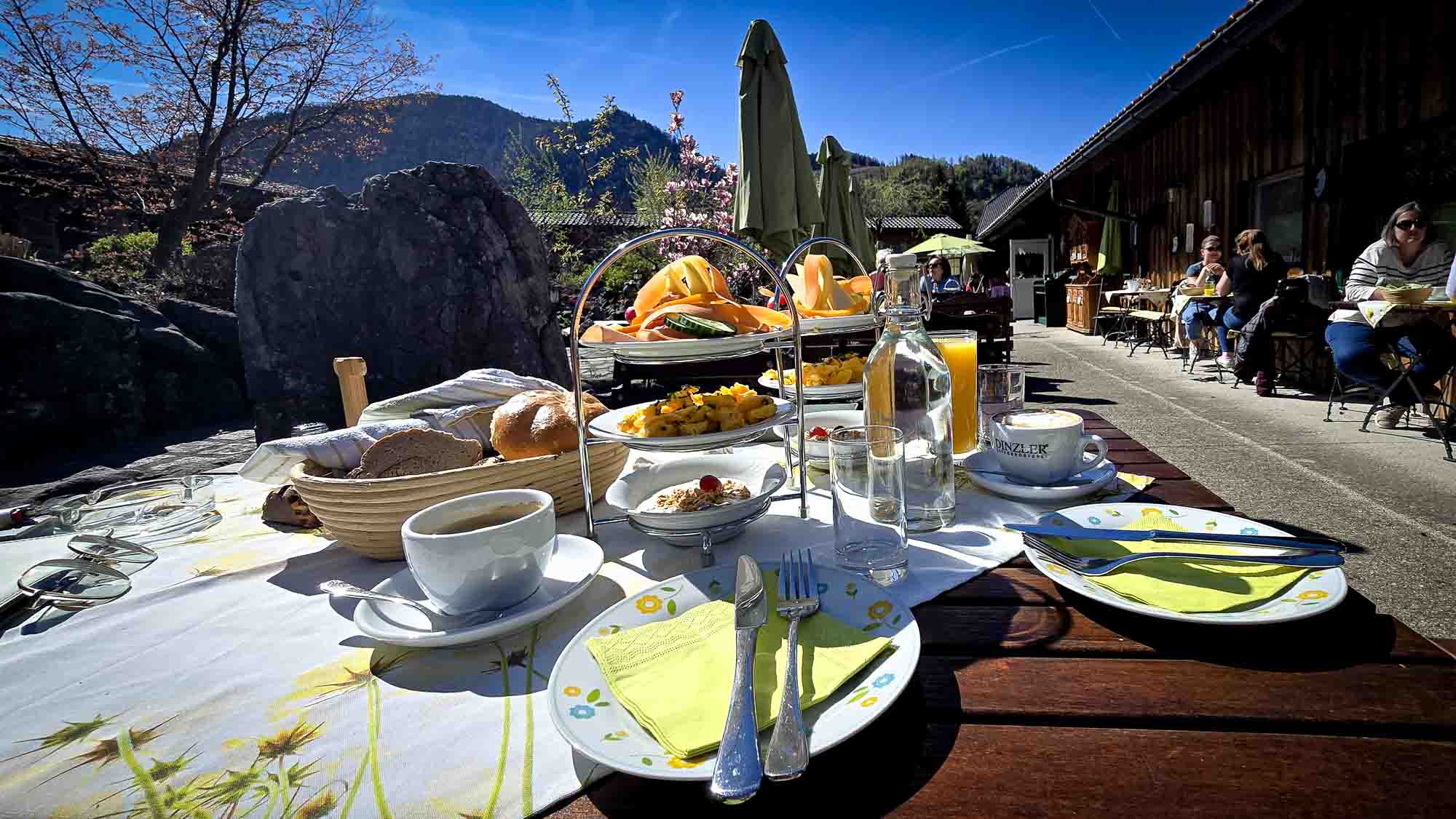 Cafétisch im Freien in Kathis Blumencafé mit Frühstücksgerichten, Obst, Kaffee und Brot vor einer sonnigen Bergkulisse. © Melanie Deisl
