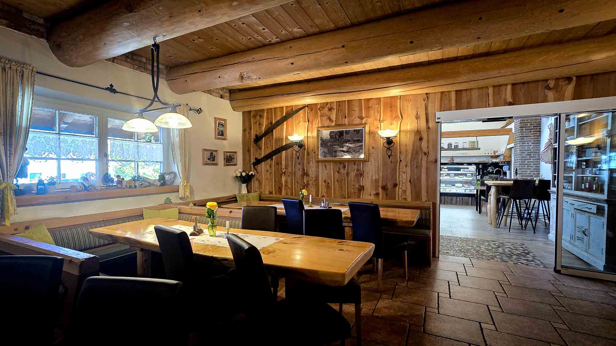 Gem&uuml;tlicher, rustikaler Essbereich in Kathis Blumencaf&eacute; mit Holztischen, schwarzen St&uuml;hlen und warmer Beleuchtung am Fenster.