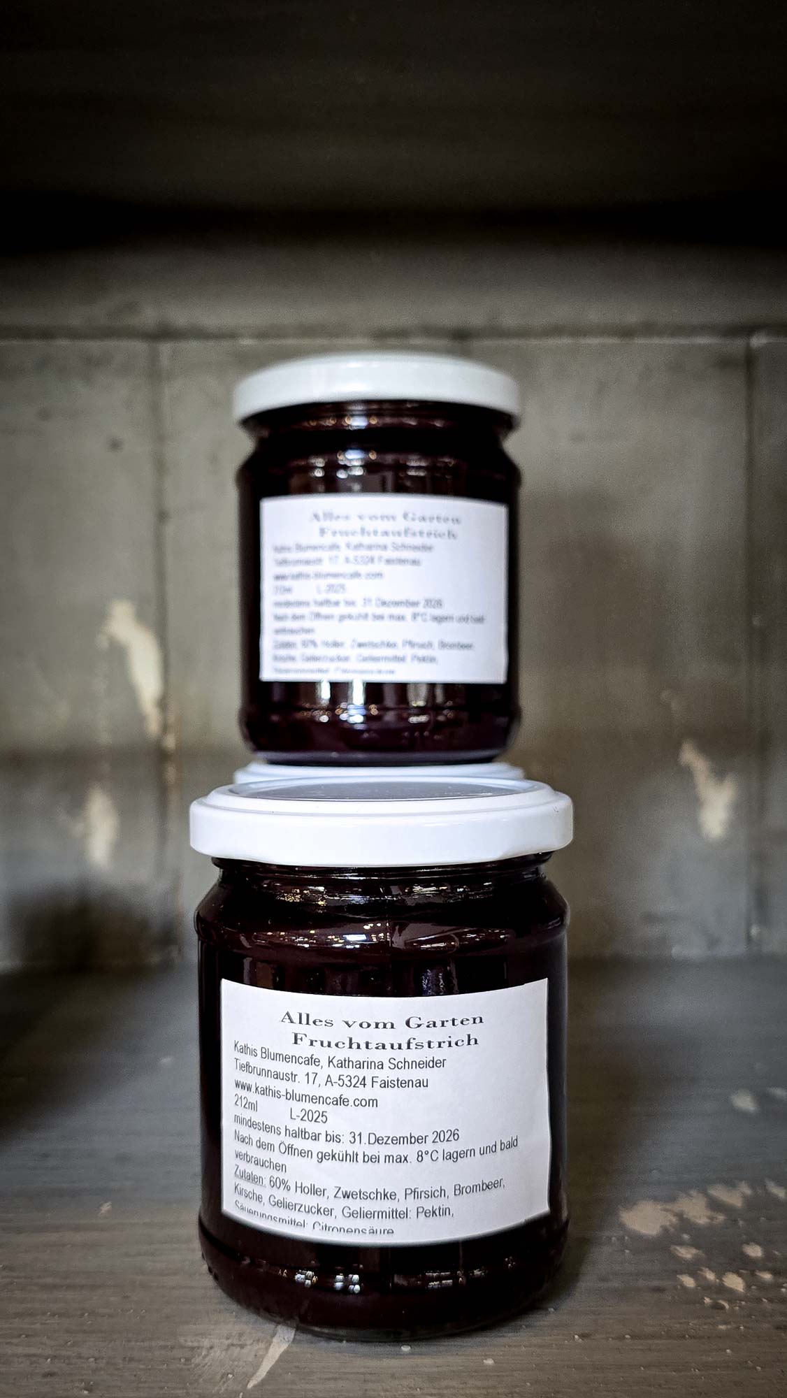 Zwei Gl&auml;ser mit selbstgemachter Marmelade von Kathis Blumencaf&eacute;, mit wei&szlig;en Deckeln und Etiketten, gestapelt auf einem Holzregal.