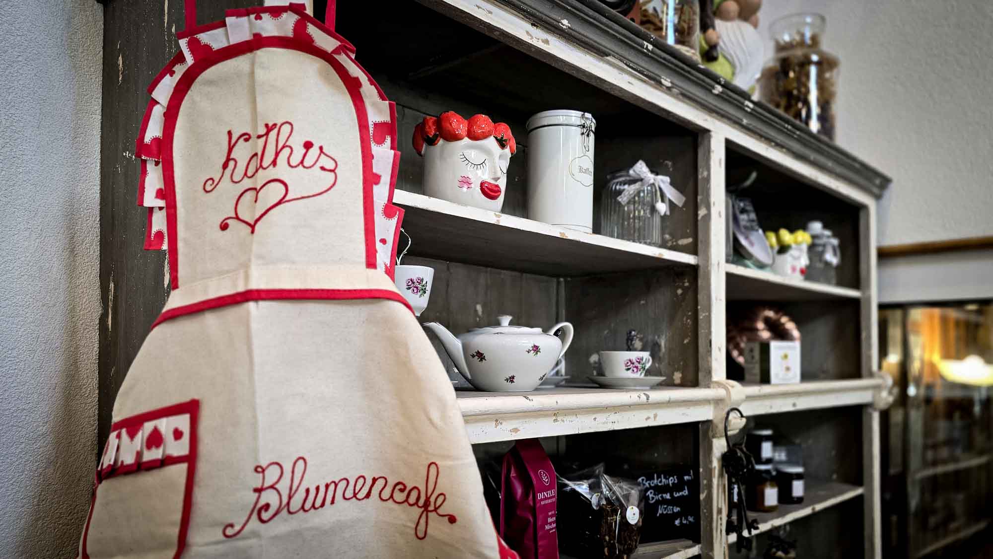 Eine Sch&uuml;rze mit der Aufschrift "Kathis Blumencaf&eacute;" h&auml;ngt neben einem Regal, das mit Gl&auml;sern, Tassen und Caf&eacute;-Deko gef&uuml;llt ist.