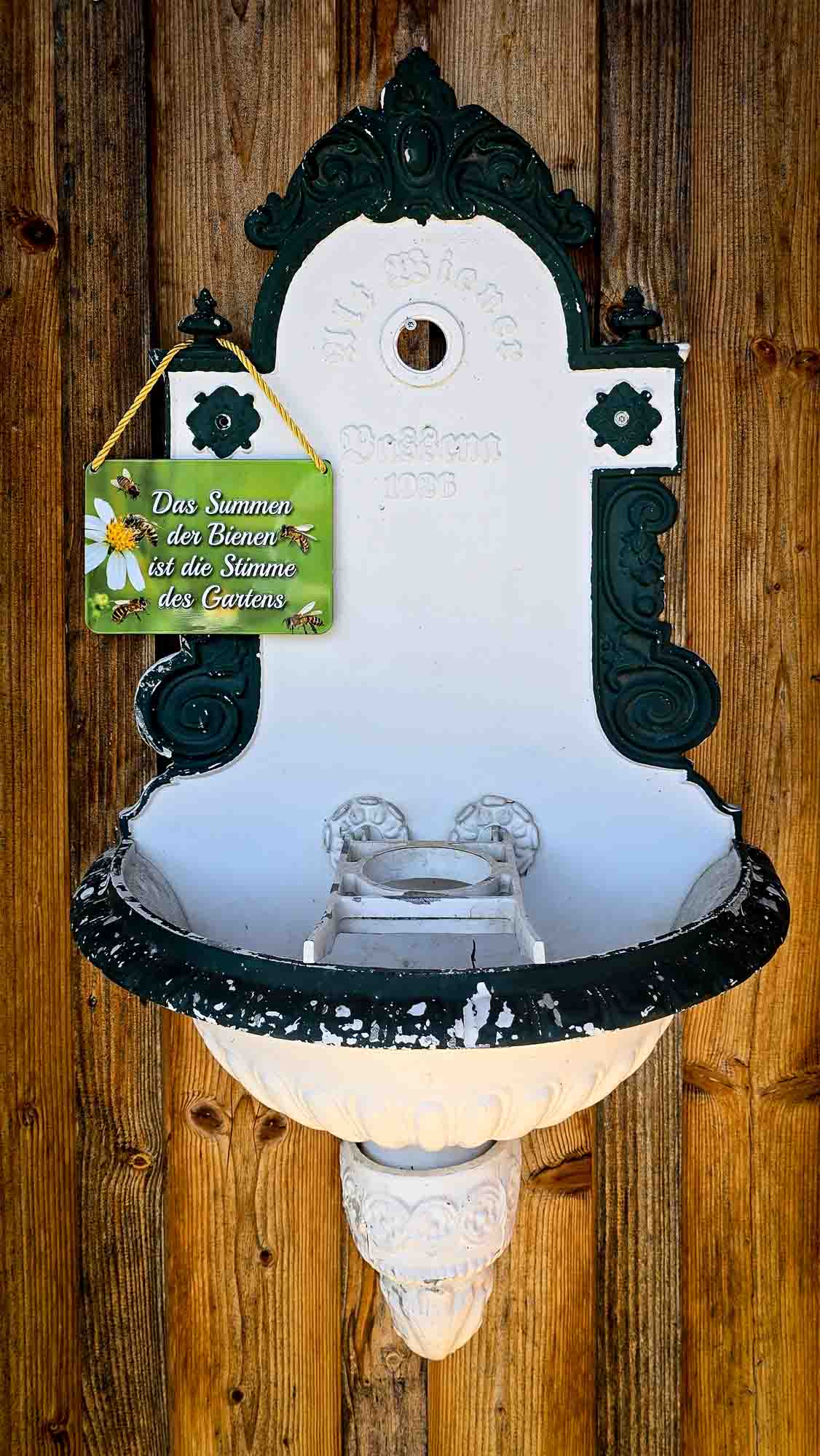 Verschn&ouml;rkelter Vintage-Wandbrunnen in Kathis Blumencaf&eacute;, mit deutschem Schild und Blumendekoration an der Holzwand.