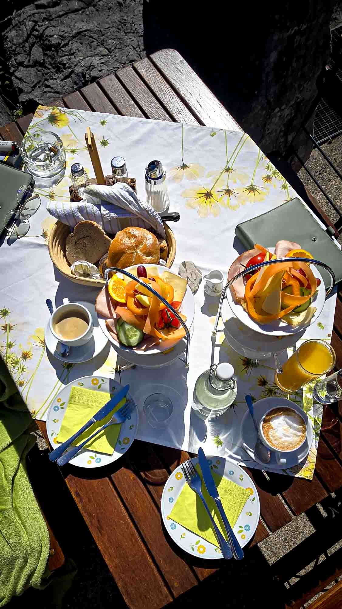 Der Fr&uuml;hst&uuml;ckstisch in Kathis Blumencaf&eacute; ist im Freien gedeckt, mit Obstschalen, Kaffee, Saft und Brot auf einer Blumentischdecke.