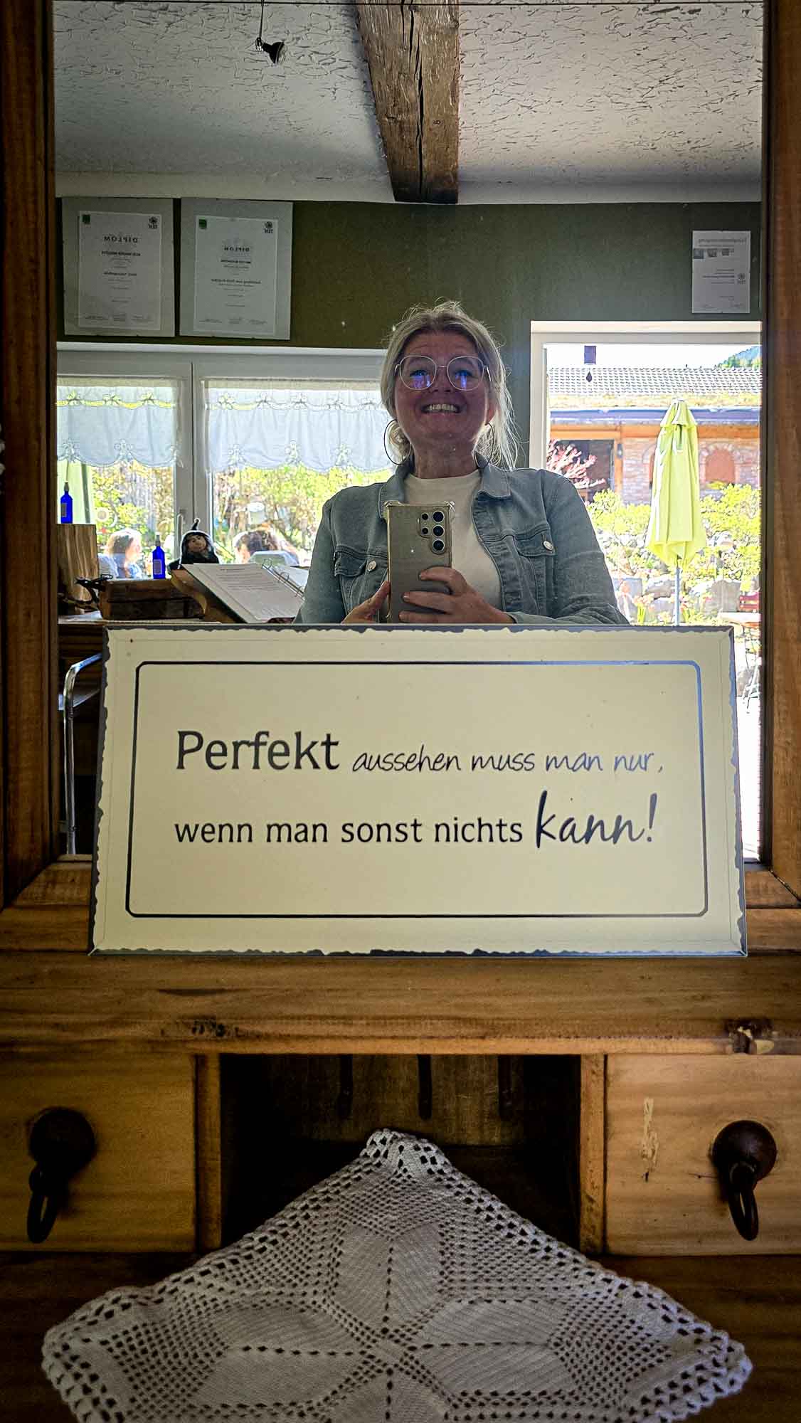 Eine Person macht ein Spiegel-Selfie in Kathis Blumencaf&eacute; und h&auml;lt ein Schild mit einem deutschen Text &uuml;ber perfektes Aussehen.