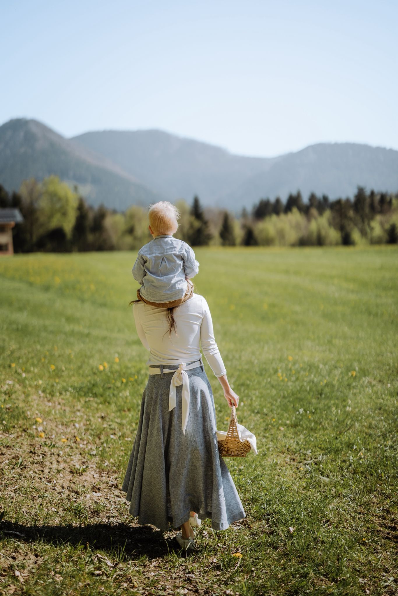 Frau, die ein kleines Kind auf den Schultern tr&auml;gt, w&auml;hrend sie durch ein grasbewachsenes Feld in Richtung Berge geht.