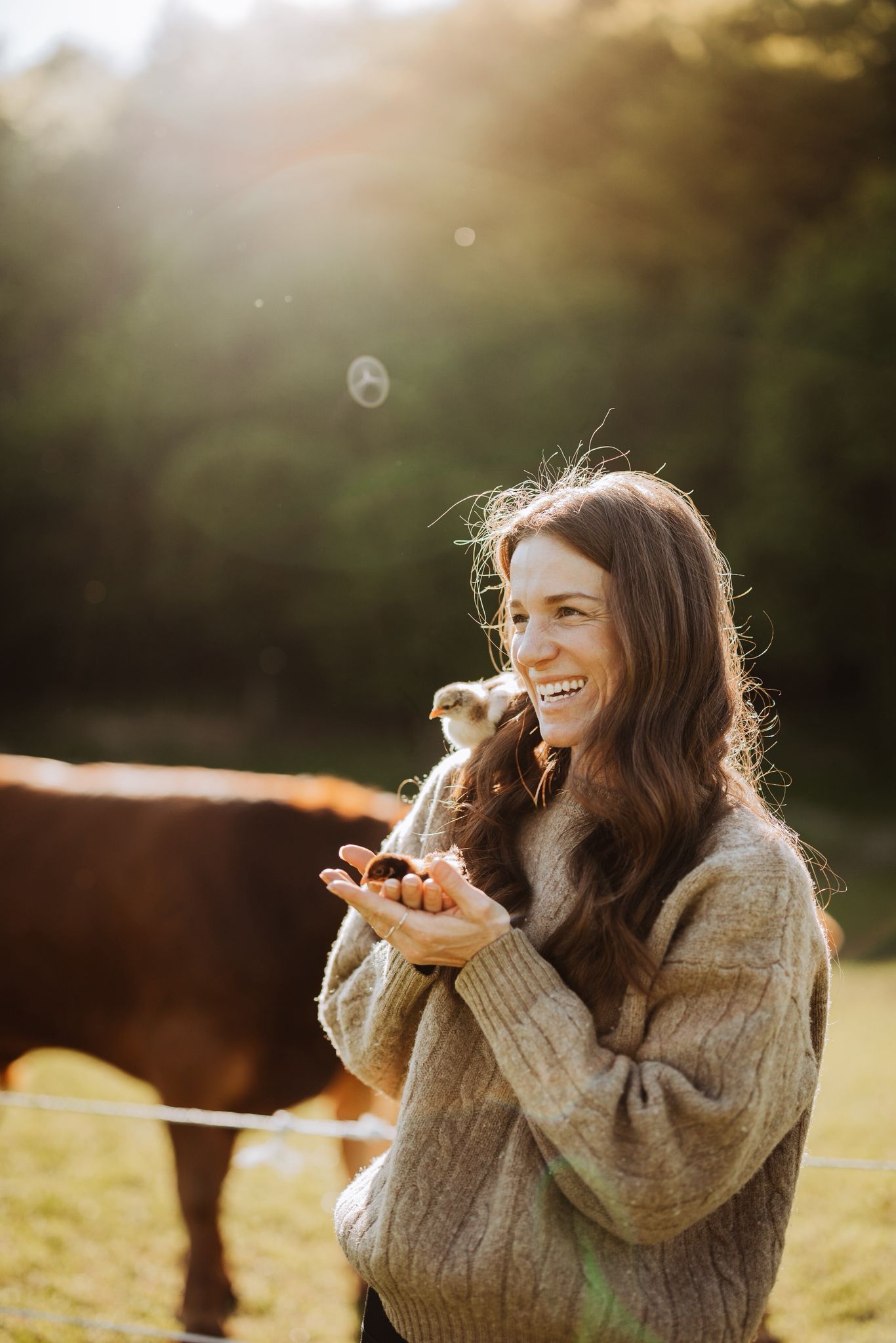 Eine l&auml;chelnde Frau im Freien h&auml;lt ein K&uuml;ken in den H&auml;nden, im Hintergrund sind eine Kuh und B&auml;ume zu sehen.