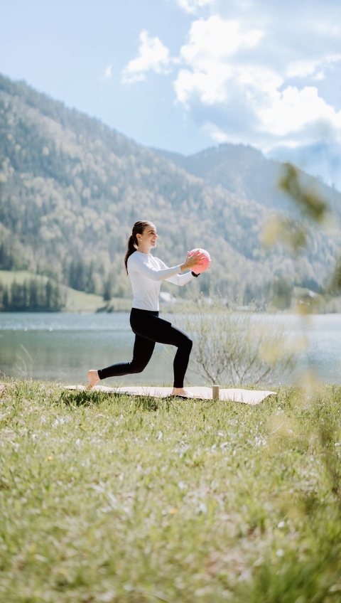 Frau macht einen Pilatesflows-Lunge mit einem rosa Ball im Freien an einem See mit Bergen im Hintergrund.