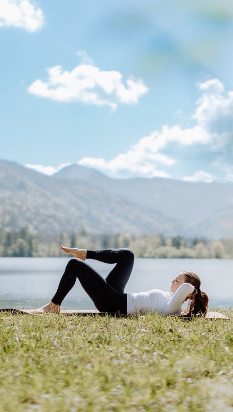 Frau macht Pilates Flows auf einer Matte im Freien an einem See, mit Bergen und blauem Himmel im Hintergrund.