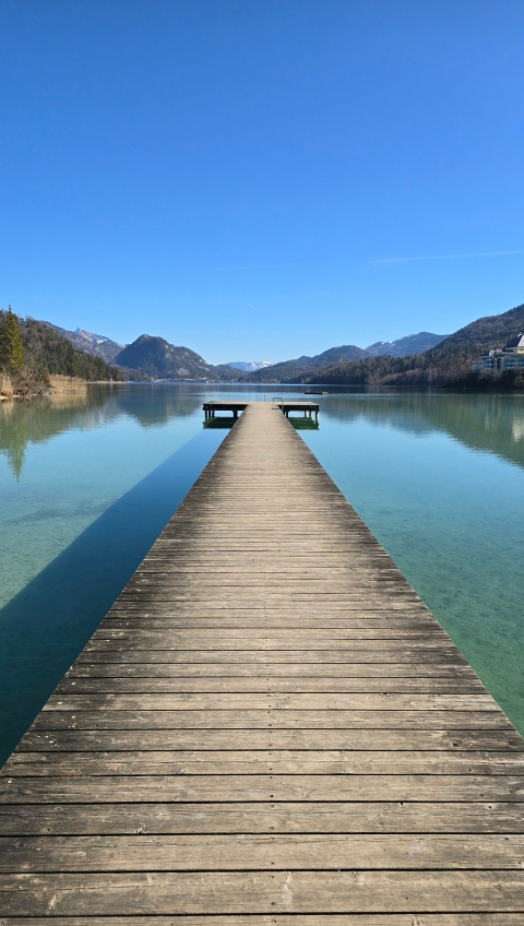 Der h&ouml;lzerne Steg erstreckt sich &uuml;ber einen klaren, ruhigen See mit Bergen und blauem Himmel im Hintergrund.