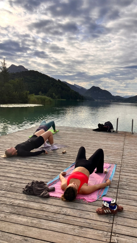 Zwei Personen trainieren auf einem h&ouml;lzernen Steg an einem malerischen See, mit Bergen und einem bew&ouml;lkten Himmel im Hintergrund.