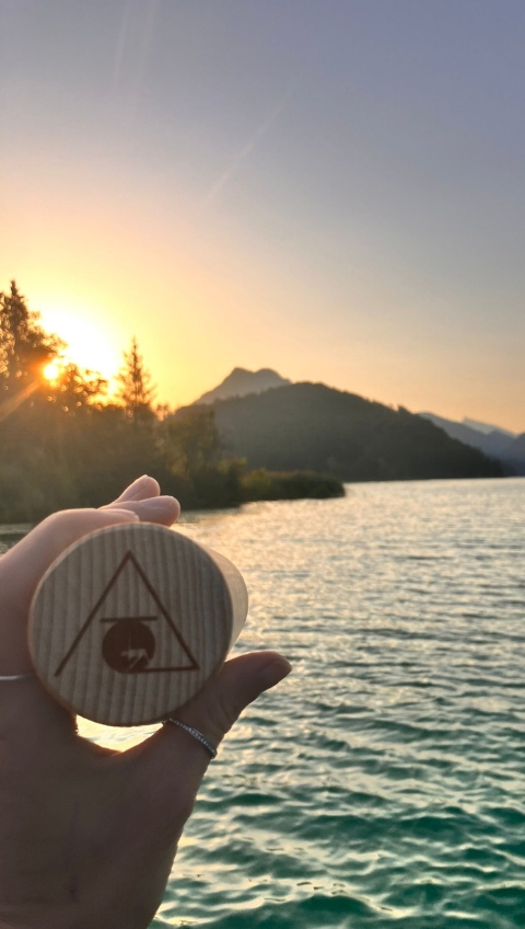 Eine Hand h&auml;lt ein rundes Holzobjekt an einem See bei Sonnenuntergang, mit Bergen und B&auml;umen im Hintergrund.