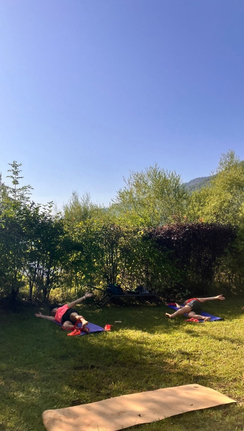 Zwei Menschen liegen auf Yogamatten in einem grasbewachsenen Au&szlig;enbereich und dehnen sich unter einem klaren blauen Himmel.