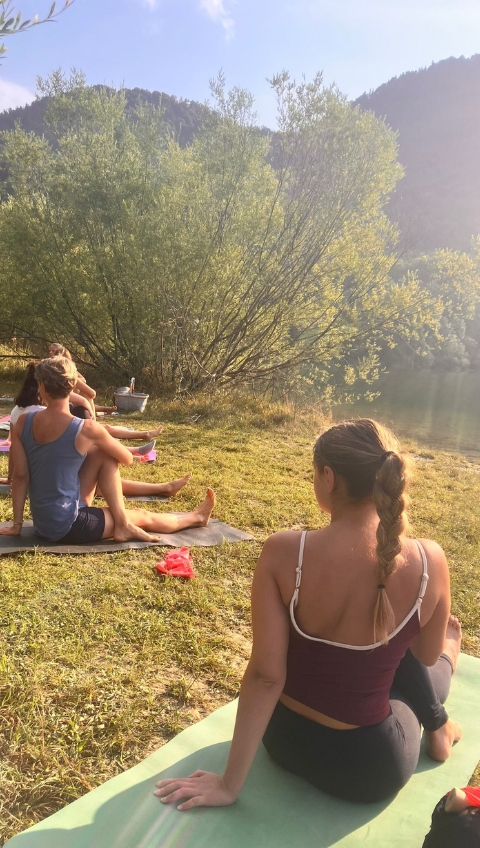 Menschen &uuml;ben Yoga im Freien in der N&auml;he eines Sees, auf Matten sitzend, mit B&auml;umen und H&uuml;geln im Hintergrund.