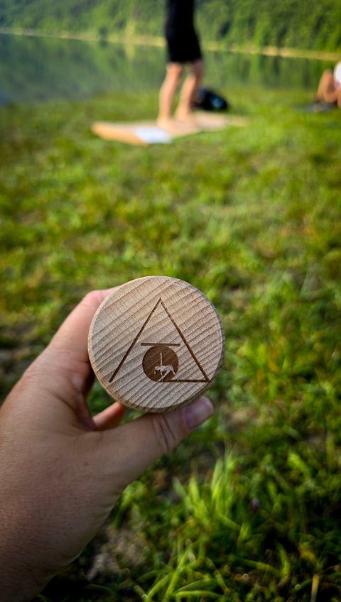 Eine Hand h&auml;lt ein rundes Holzobjekt mit einem Dreieck und einem Berg-Logo im Freien in der N&auml;he eines Sees.