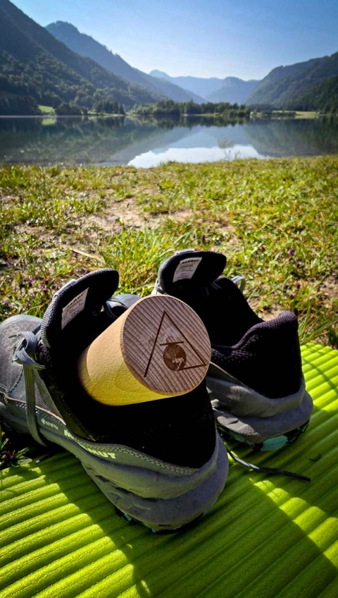 Laufschuhe und eine Holzrolle auf einer Matte an einem See, Berge und B&auml;ume im Hintergrund unter einem klaren Himmel.