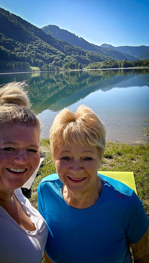 Zwei Frauen l&auml;cheln an einem See mit Bergen und B&auml;umen im Hintergrund an einem sonnigen Tag.