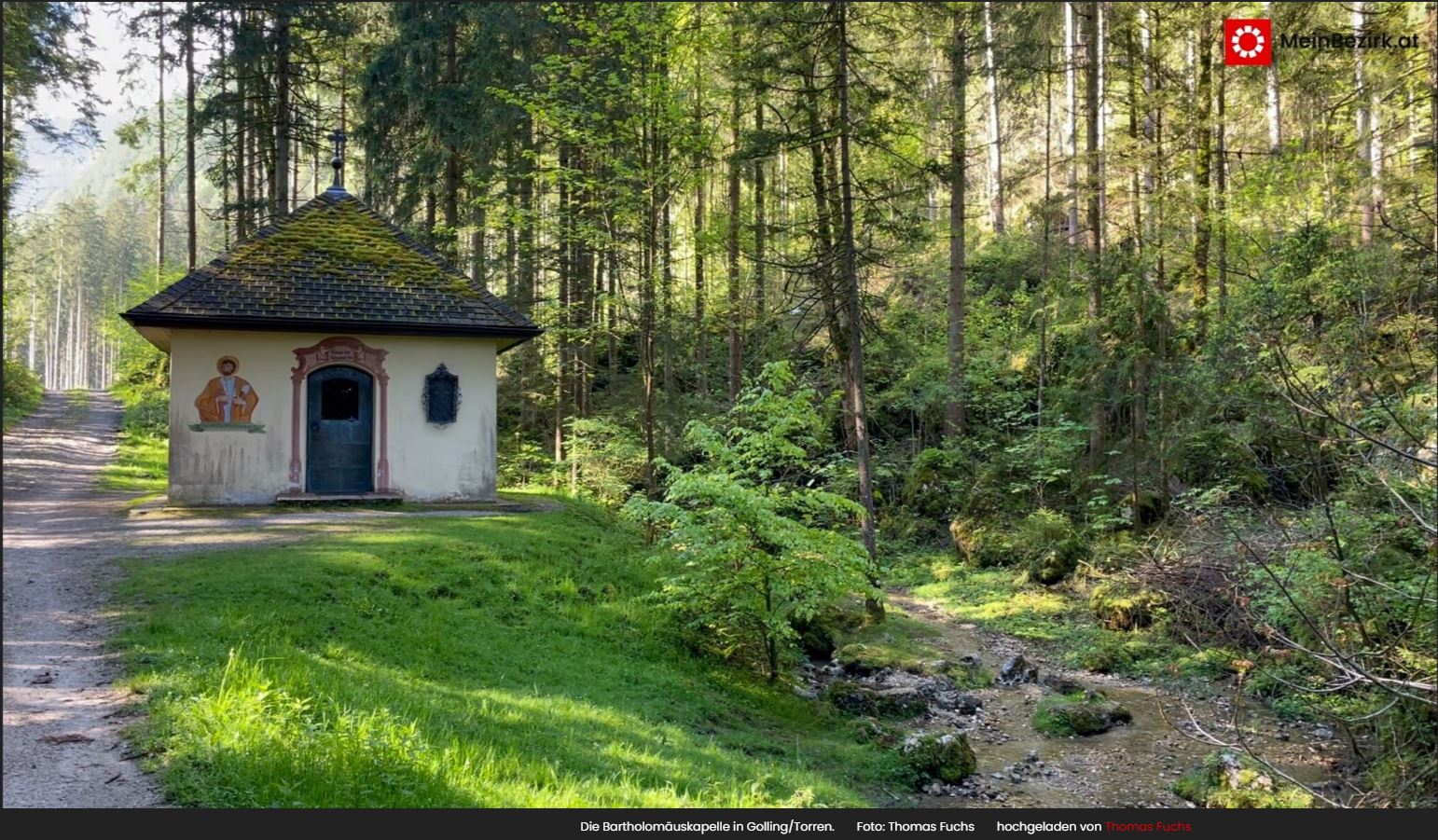 Kleine Kapelle an einem Waldweg, umgeben von hohen B&auml;umen und einem Bach, der durch gr&uuml;nes Gestr&uuml;pp flie&szlig;t.