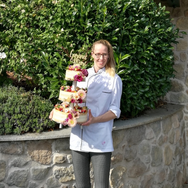 Eine Frau im Kochmantel h&auml;lt eine Blumentorte im Freien bei Kathis Blumencaf&eacute;, an einer Steinmauer und im Gr&uuml;nen.