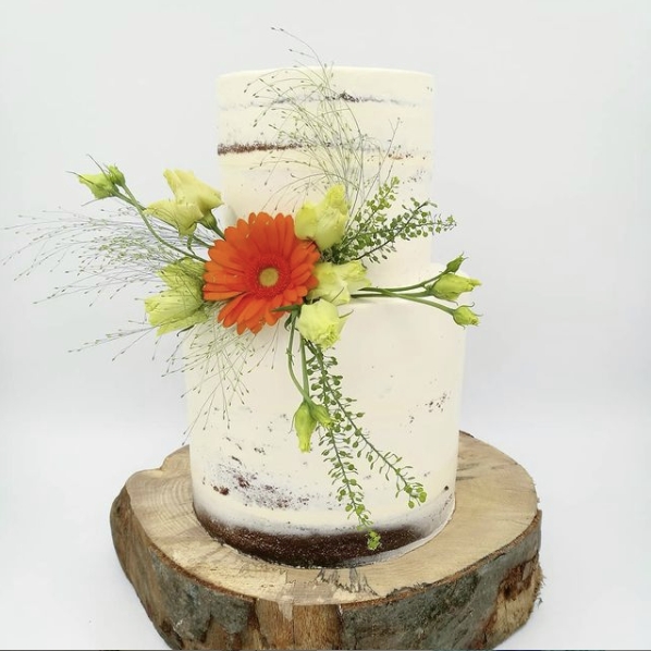 Minimalistische wei&szlig;e Torte von Kathis Blumencaf&eacute; mit orangefarbenen und gelben Blumen auf einem rustikalen Holzst&auml;nder.