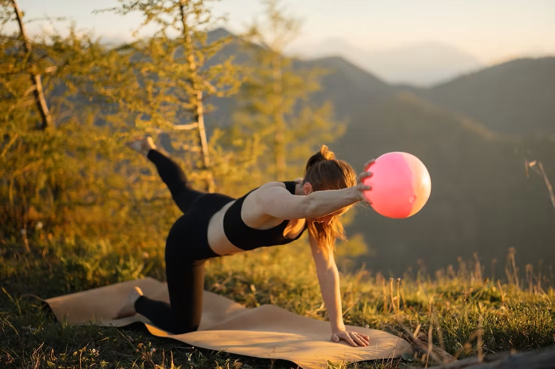 Frau macht Yoga auf einer Matte im Freien, balanciert auf einem Knie und einer Hand, h&auml;lt einen rosa Ball, Berge im Hintergrund.