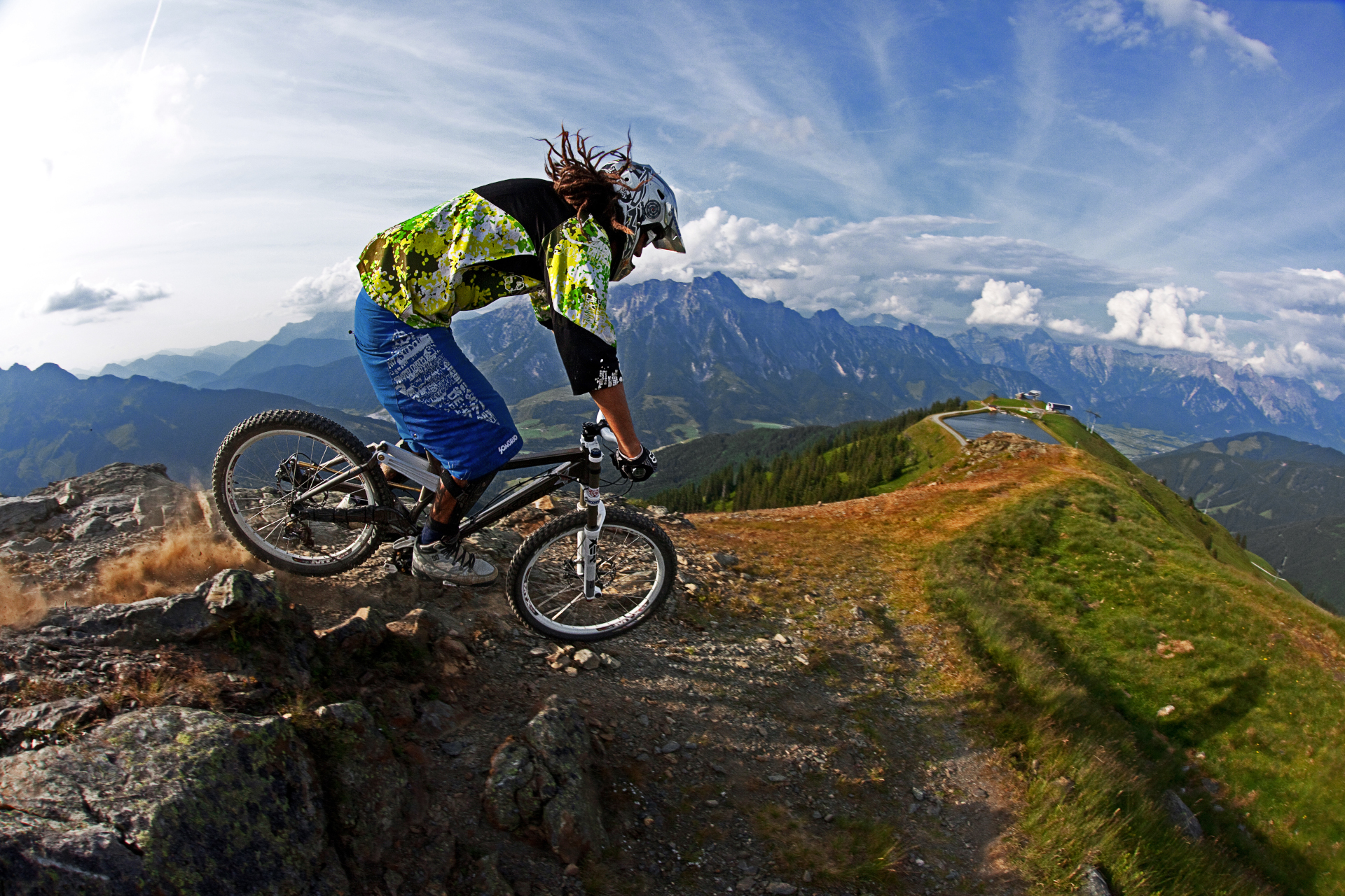 Mountainbiker f&auml;hrt an einem sonnigen Tag einen felsigen Weg hinunter, mit Bergen und blauem Himmel im Hintergrund.