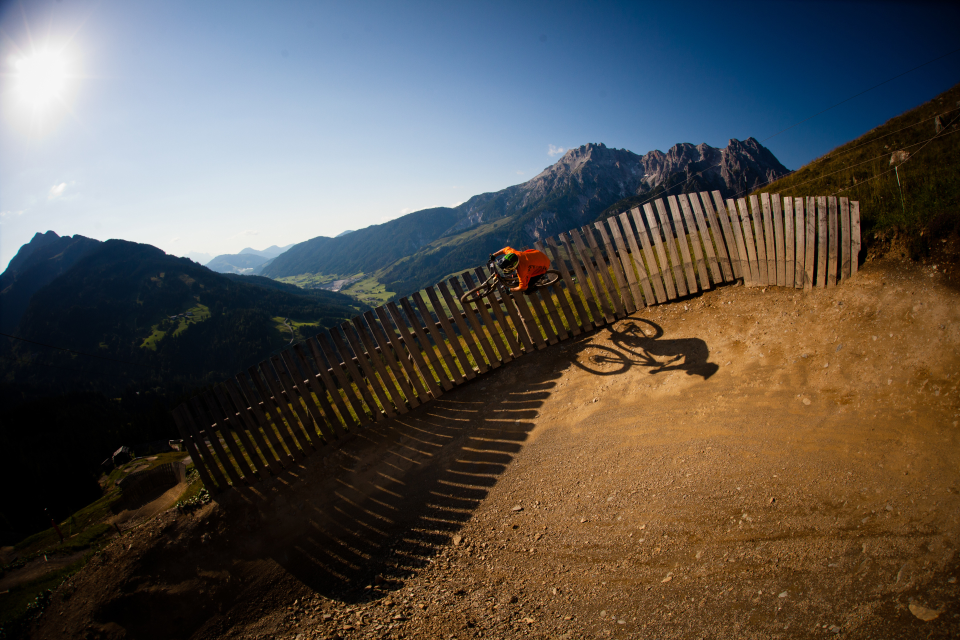 Ein Mountainbiker f&auml;hrt eine gebogene Holzrampe auf einem unbefestigten Weg mit Bergen im Hintergrund hinauf.