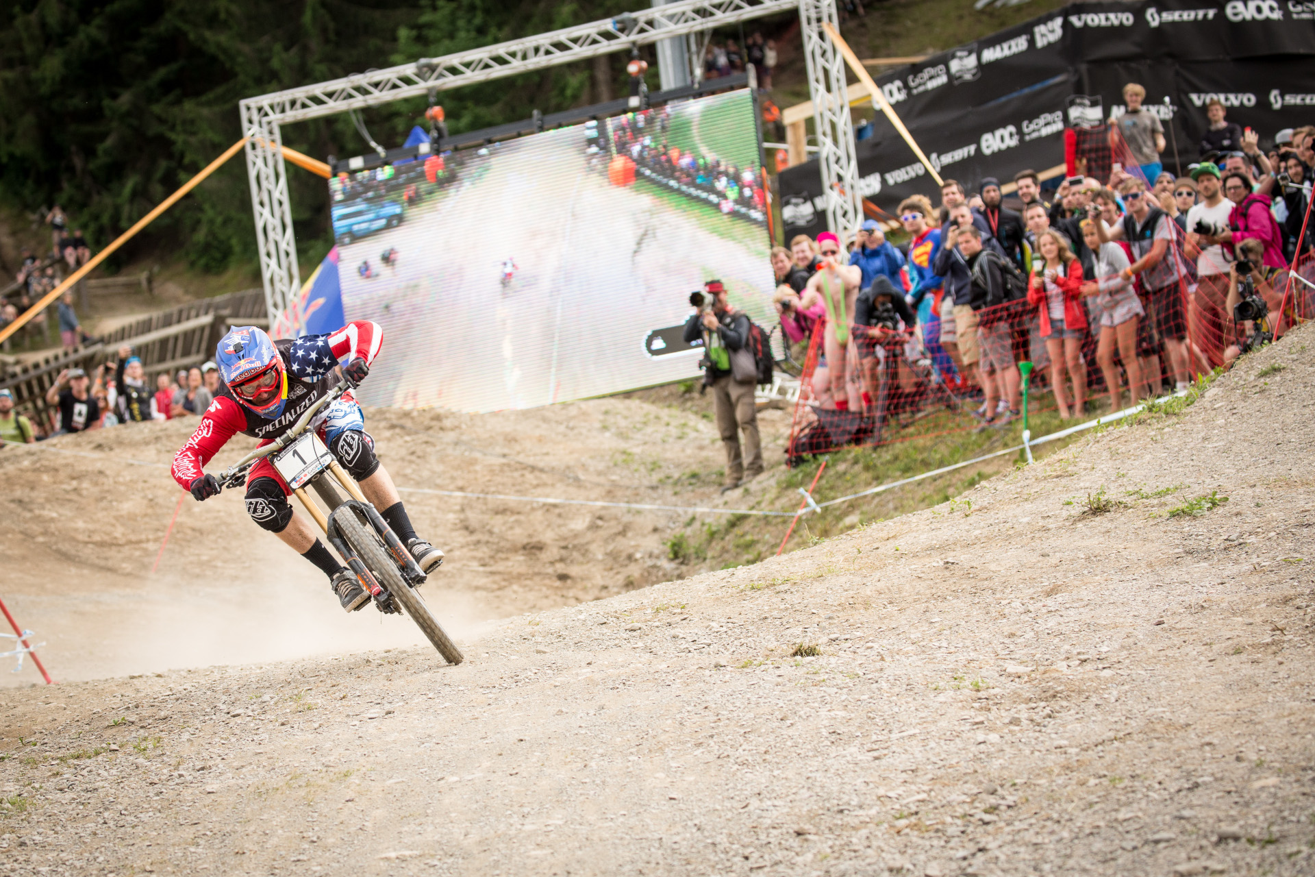 Ein Mountainbiker rast bergab, w&auml;hrend die Zuschauer bei einer Veranstaltung im Freien vor einem gro&szlig;en Bildschirm zusehen und Fotos machen.