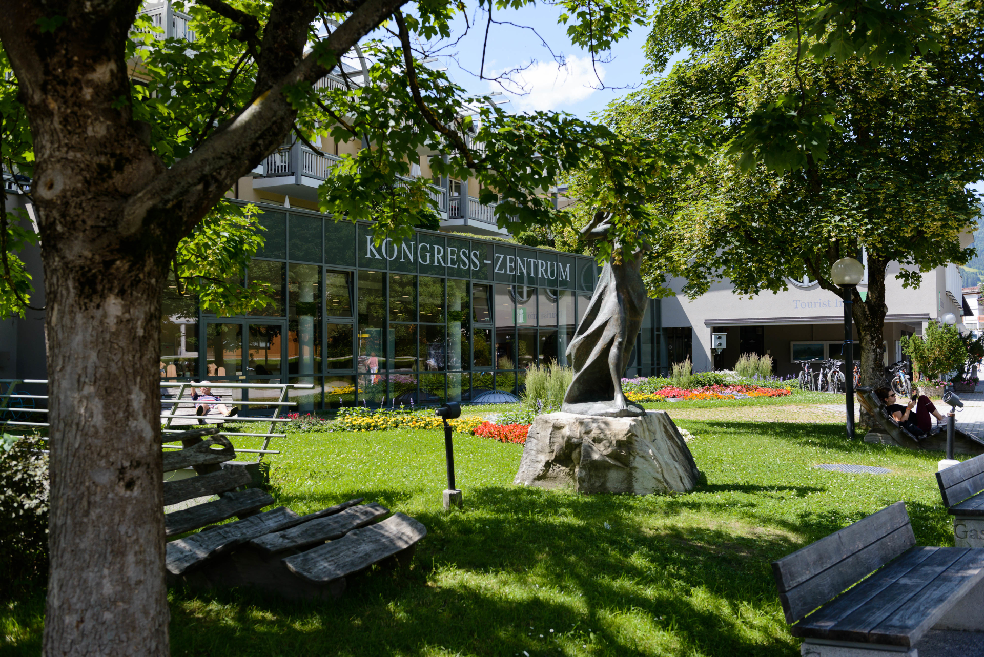 Ein baumbeschatteter Park mit B&auml;nken, einer modernen Skulptur und einem Geb&auml;ude mit der Aufschrift Kongress-Zentrum.