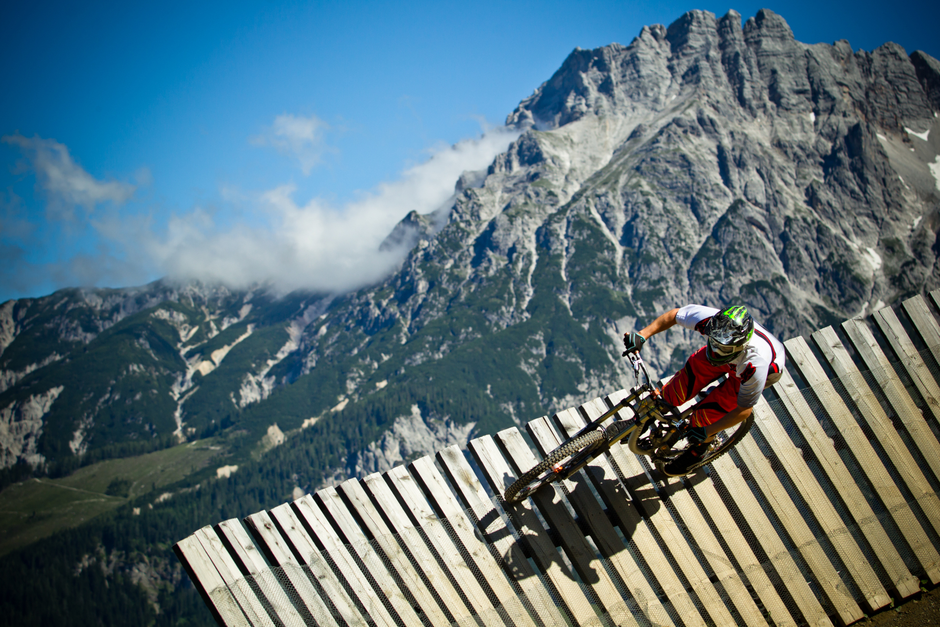 Mountainbiker f&auml;hrt auf einer h&ouml;lzernen Rampe mit einem hoch aufragenden felsigen Berg im Hintergrund unter einem blauen Himmel.