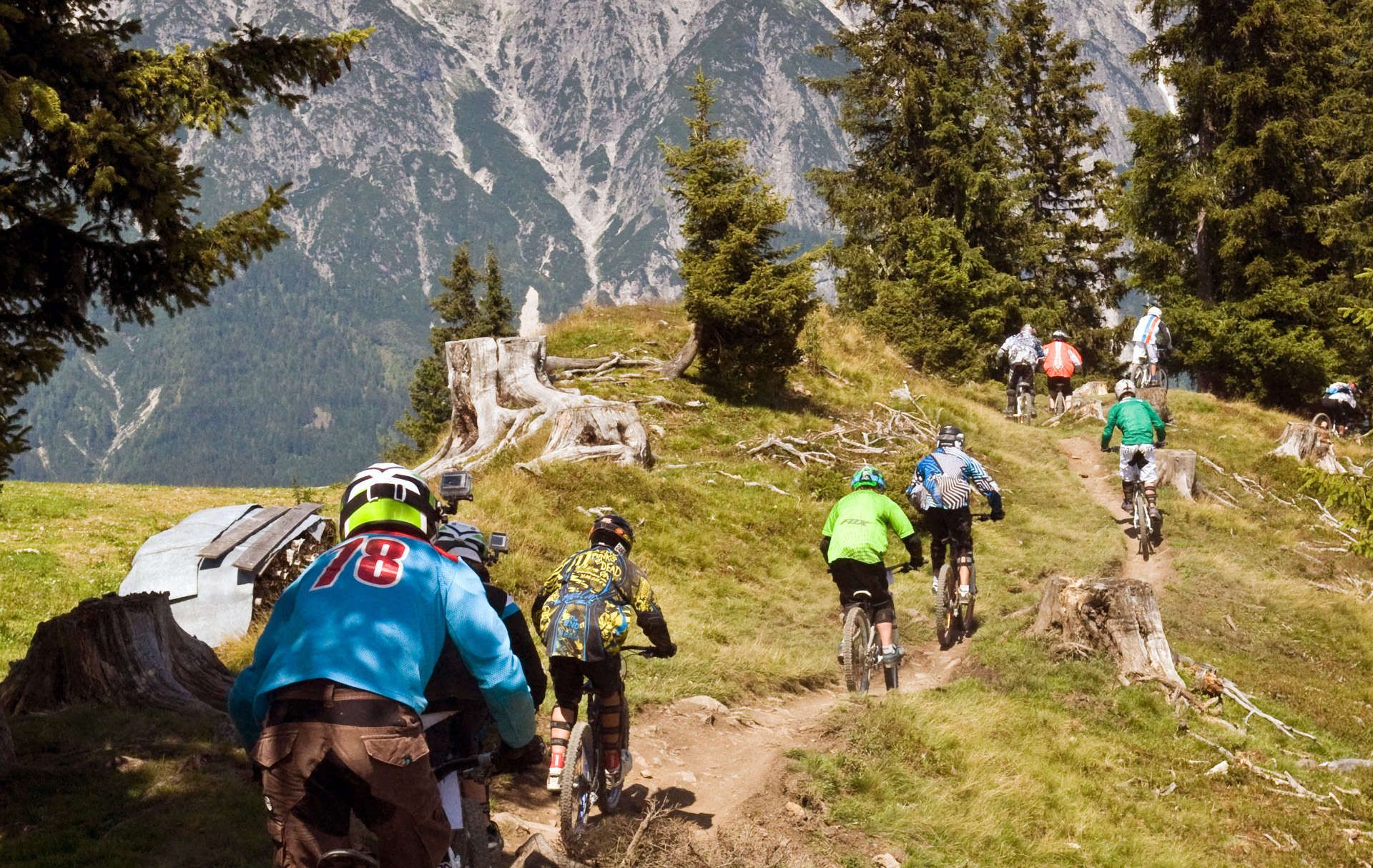 Mehrere Mountainbiker fahren auf einem wilden Waldweg mit Bergen im Hintergrund.
