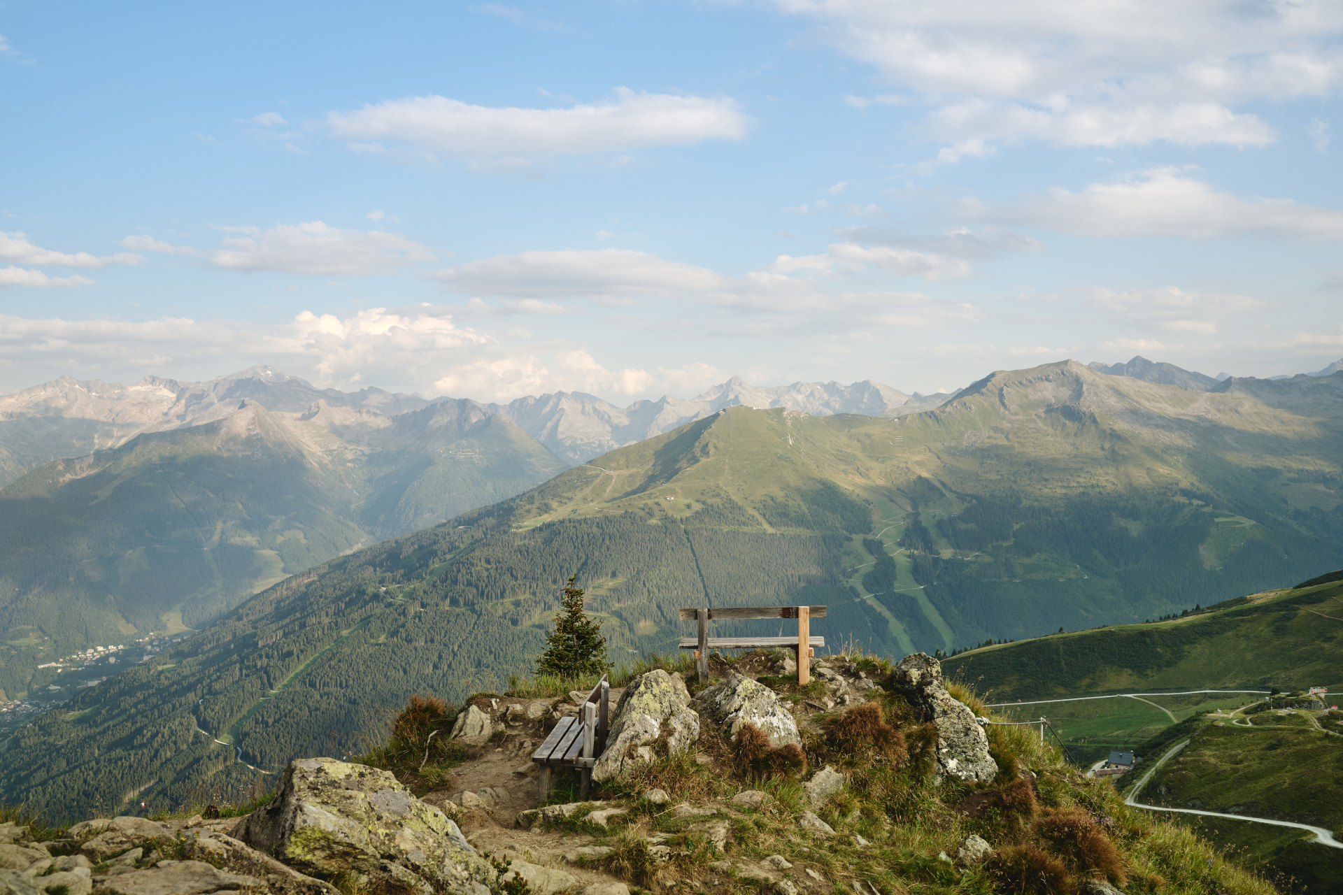Holzbank auf einem felsigen Hügel mit Blick auf grüne Berge und einen blauen Himmel mit vereinzelten Wolken.
