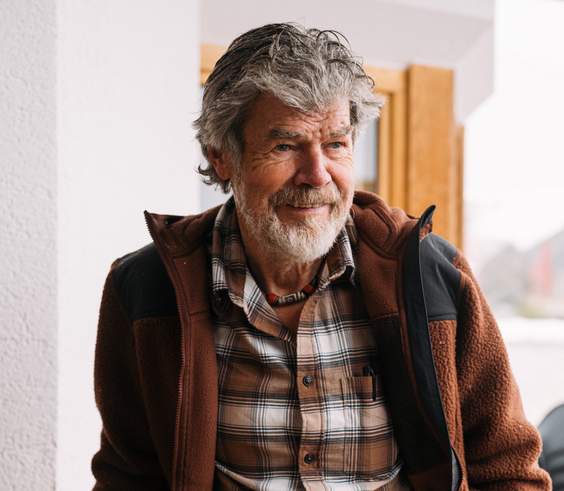Älterer Mann mit grauem Haar und Bart, der eine braune Jacke und ein kariertes Hemd trägt, lächelt im Freien in der Nähe eines Fensters.
