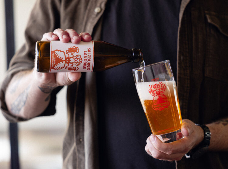 Eine Person gießt Salzburger Lagerbier aus einer Flasche in ein Glas, beide mit rotem Logo versehen.