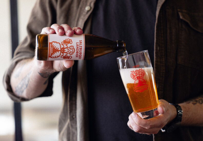 Eine Person gießt Salzburger Lagerbier aus einer Flasche in ein Glas, beide mit rotem Logo versehen.
