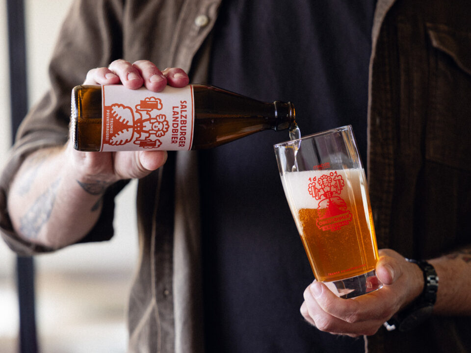 Eine Person gießt Salzburger Lagerbier aus einer Flasche in ein Glas, beide mit rotem Logo versehen.