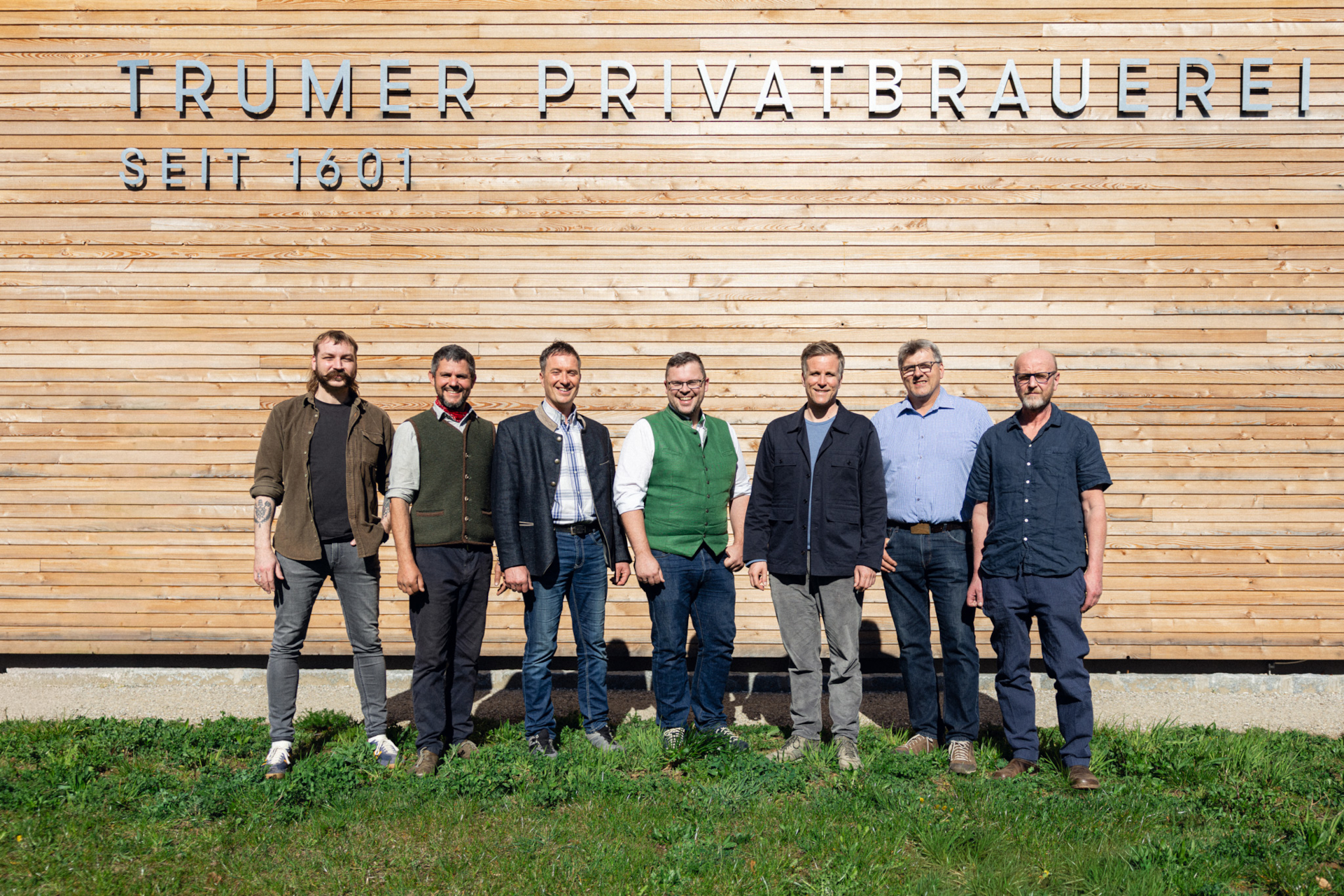 Sieben Männer stehen lächelnd vor einer Holzwand mit der Aufschrift Trumer Privatbrauerei Seit 1601.