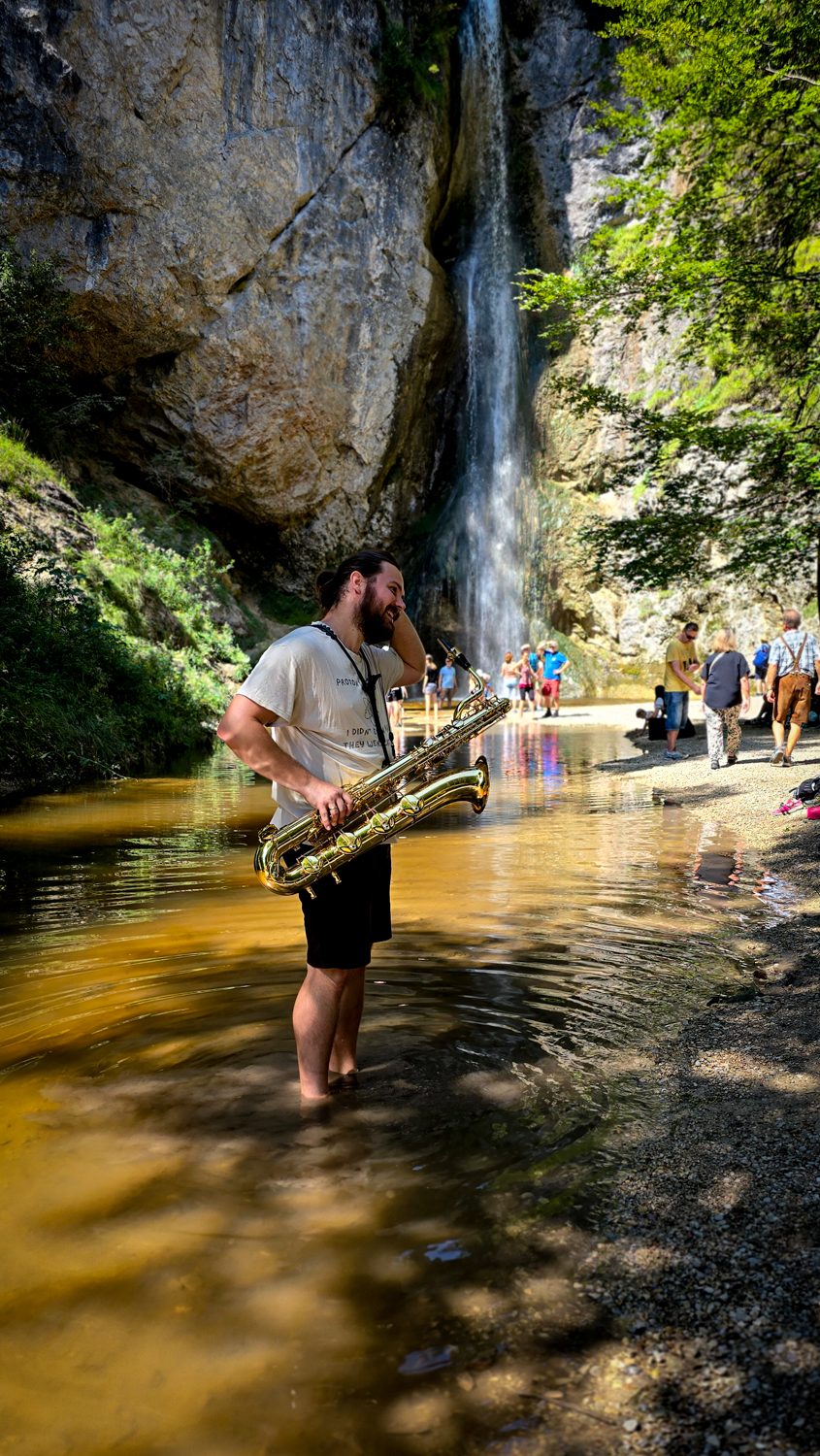 Ein Mann mit einem Saxophon steht beim M&uuml;hlenfest in Ebenau in einem Bach, an dessen Wasserfall sich Menschen versammelt haben.