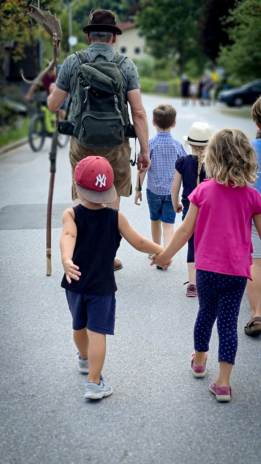 Ein &auml;lterer Mann mit einem Rucksack f&uuml;hrt Kinder, die sich an den H&auml;nden halten, zum M&uuml;hlenfest in Ebenau.