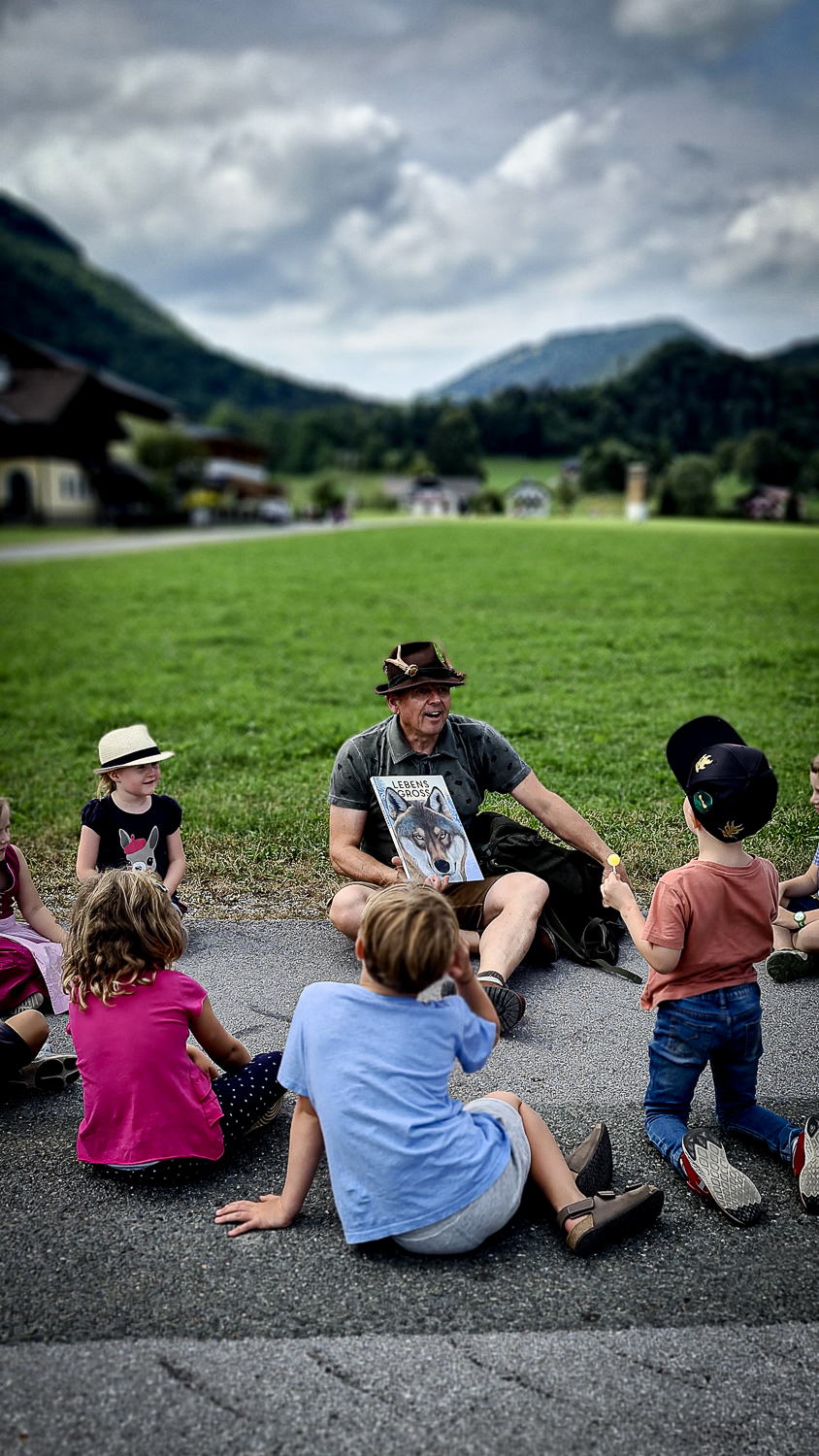 Ein Mann teilt ein Bilderbuch mit Kindern auf dem M&uuml;hlenfest in Ebenau, im Hintergrund die Berge.