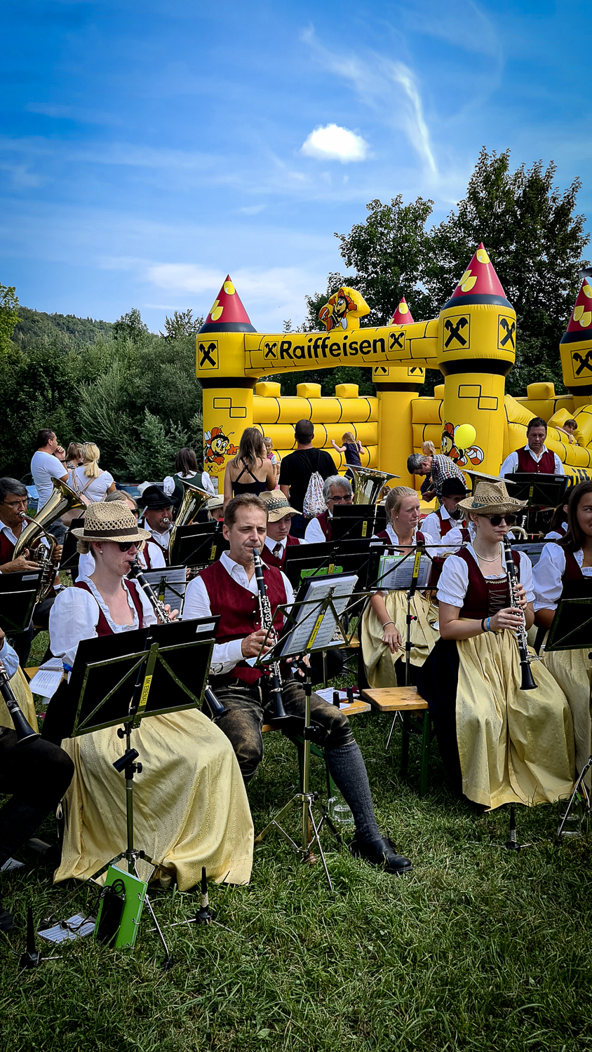 Eine Trachtenkapelle spielt beim M&uuml;hlenfest in Ebenau neben einer gelben Raiffeisen-H&uuml;pfburg.