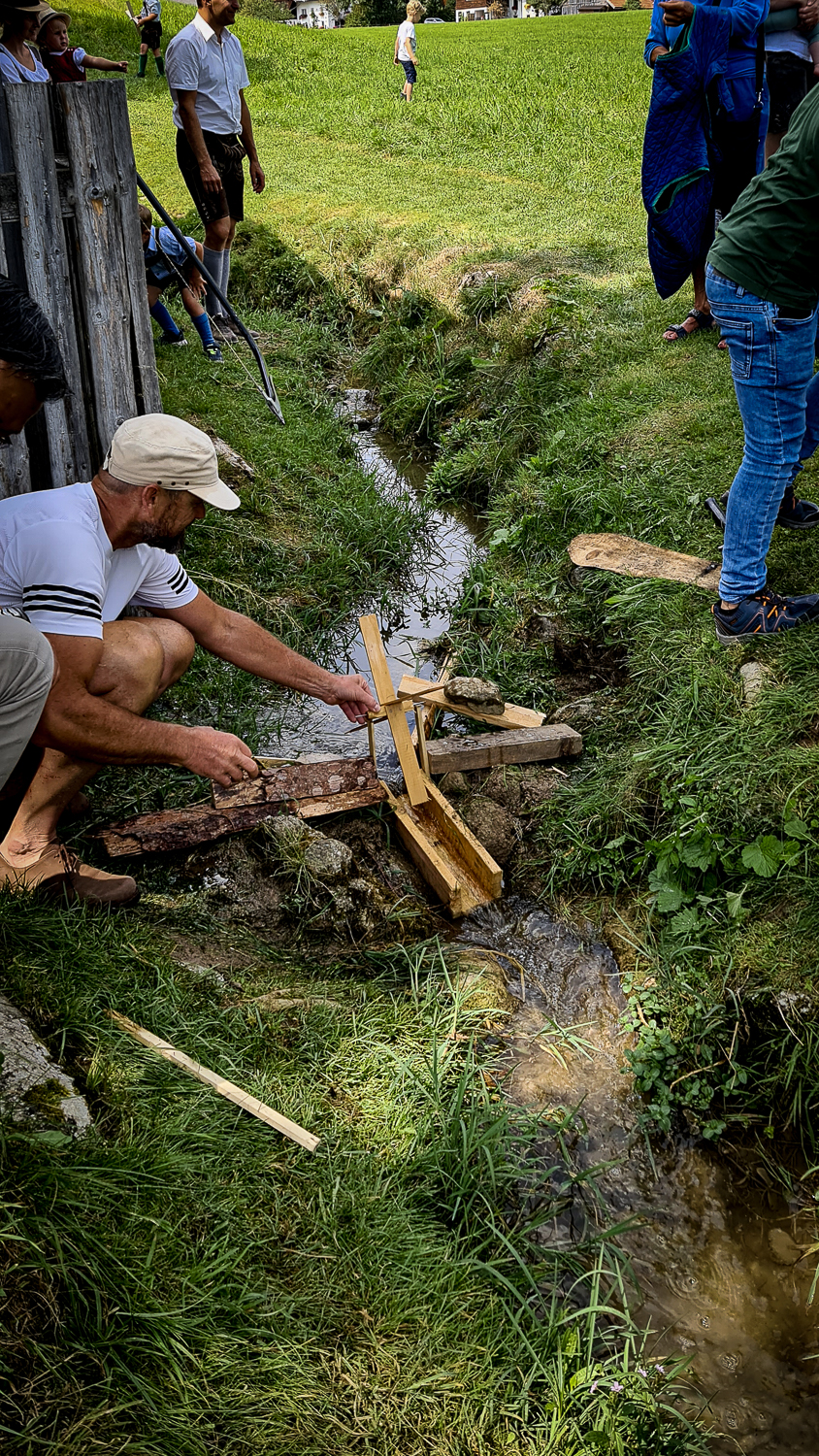 Menschen bauen beim M&uuml;hlenfest in Ebenau eine kleine h&ouml;lzerne Wasserm&uuml;hle &uuml;ber einem schmalen Bach auf einer Wiese.