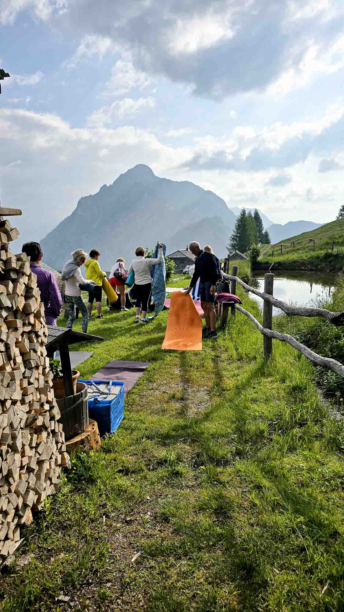 Eine Gruppe praktiziert Yoga auf Matten, die im Gras in der N&auml;he eines Sees ausgelegt sind, mit den Bergen im Hintergrund.