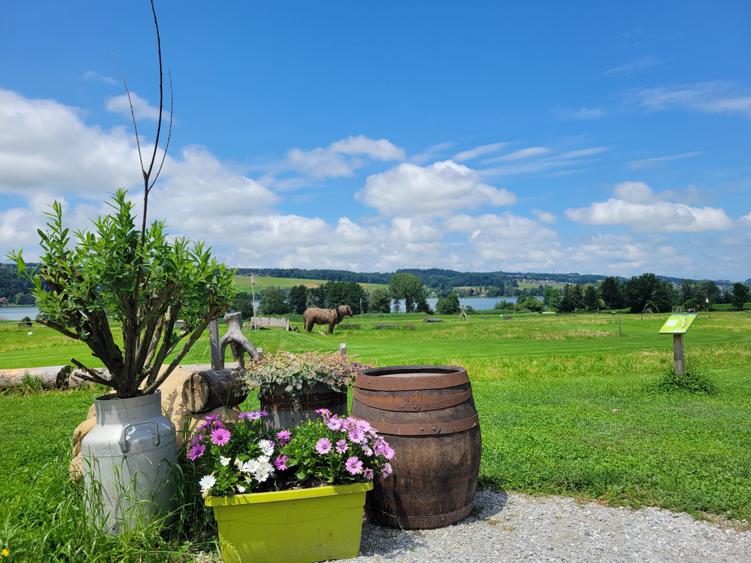 Ein Fass, ein Milchk&auml;nnchen und Blument&ouml;pfe vor einem gr&uuml;nen Feld, im Hintergrund ein Pferd und ein blauer Himmel.