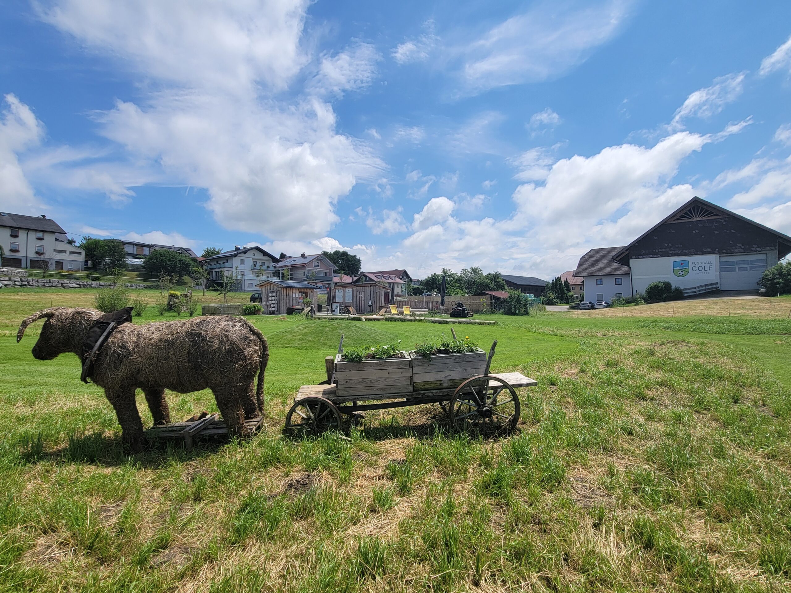 Auf einer Wiese in der N&auml;he einiger Geb&auml;ude steht unter einem blauen Himmel mit Wolken die Skulptur eines Ochsen, der einen Holzwagen zieht.