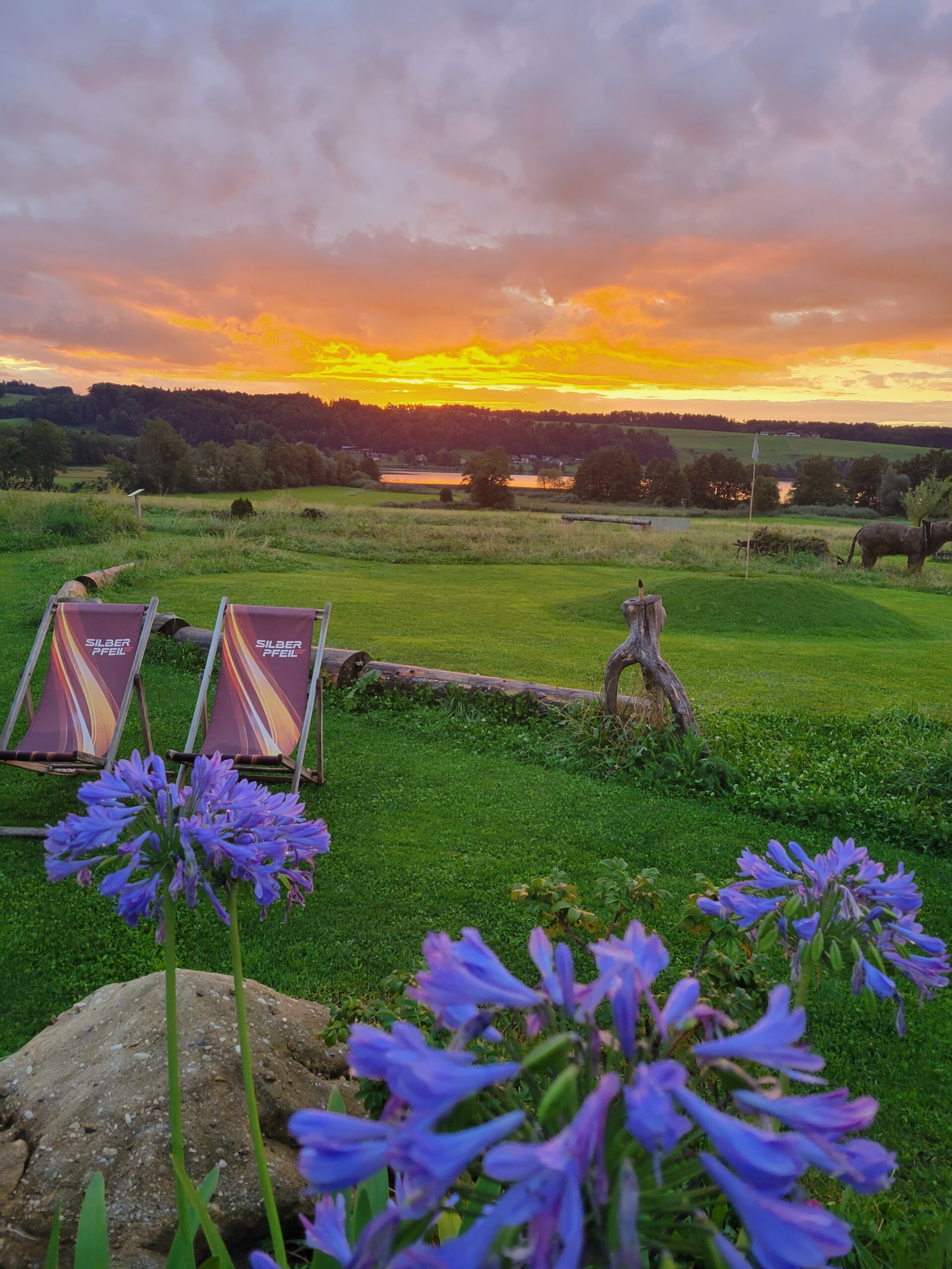 Zwei Liegest&uuml;hle auf gr&uuml;nem Rasen, davor violette Blumen, mit Blick auf eine vom Sonnenuntergang beleuchtete Landschaft und einen See.